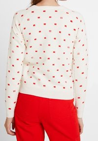 Weißer Pullover mit strukturierter Strickoptik, mit kleinen roten Flaggenmustern. Rippbündchen und Saum. Ein Paar rote Hosen darunter getragen.
