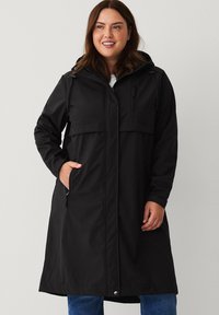 Sort vandtæt jakke med hætte, frontlynlås, to sidelommer og en brystlomme. Lange ærmer og knælangt design.