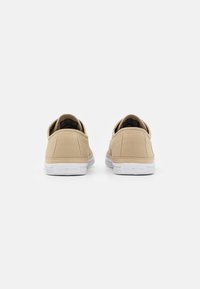 Un par de zapatillas de lona beige vistas desde atrás con suelas blancas sobre un fondo blanco sencillo.