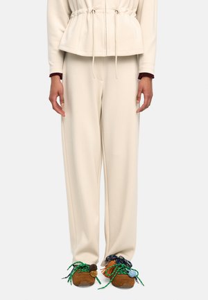 Kremowe spodnie o szerokich nogawkach w połączeniu z brązowymi trampkami z zielonymi sznurowadłami oraz ozdobnymi, wielokolorowymi frędzlami i pomponami.