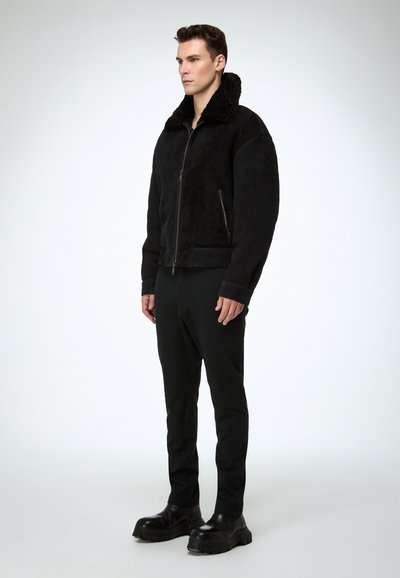 Blouson aviateur en daim noir avec col en fausse fourrure texturée, fermeture éclair frontale et poches latérales. Associé à un pantalon noir sur mesure et des chaussures massives.