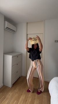 Άτομο με ριγωτό παντελόνι και μαύρο πουκάμισο κρατά κίτρινη τσάντα πάνω από το κεφάλι, όρθιο σε υπνοδωμάτιο με λευκά έπιπλα και ξύλινο πάτωμα.