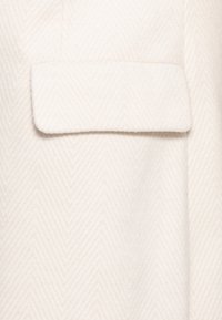 Wit stof met visgraatmotief en een zachte textuur. Voorzien van een enkele klepzak aan de linkerkant, wat detail aan het ontwerp toevoegt.