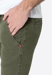 Olive green pants made of textured fabric, featuring a side pocket with a red accent button and a classic fit design.