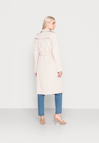 Hellbeiger Trenchcoat mit breitem Kragen und taillierter Taille. Der Mantel hat lange Ärmel und reicht bis unter das Knie, kombiniert mit blauen Jeans.