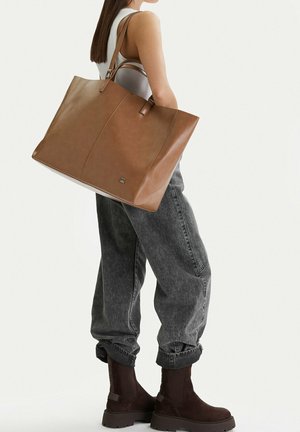 Kobieta niosąca dużą brązową skórzaną torbę na ramię, ubrana w szare jeansy, biały top bez rękawów i brązowe botki na jednolitym tle.