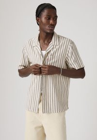 Short-sleeve, button-up shirt with beige fabric and dark striped pattern, featuring a chest pocket and lightweight texture.