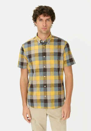 Ung mand iført en kortærmet, ternet skjorte i gult, blåt og brunt og beige cargobukser, stående mod en hvid baggrund.