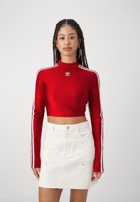 Κόκκινο μακρυμάνικο crop top με ψηλό λαιμό και λευκό λογότυπο Adidas, με λευκές πλευρικές ρίγες. Συνδυάζεται με λευκή τζιν φούστα.