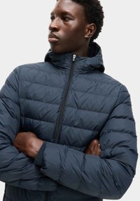 Giacca imbottita blu navy con colletto alto, chiusura con zip e pannelli cuciti. Presenta un cappuccio e polsini elasticizzati. Texture morbida e liscia.
