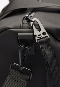 Sac de voyage noir avec une texture lisse, tissu résistant, fermeture éclair en métal et clip en plastique avec une sangle noire réglable.