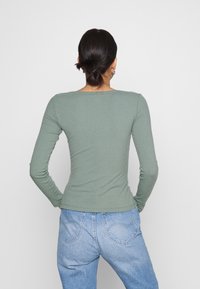 Langærmet, tætsiddende ribbet top i salviegrøn. Rund hals, glat tekstur. Sættes sammen med højtaljede lyseblå jeans med hjerteformede lommer.