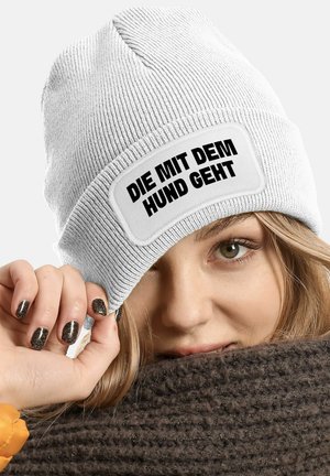 White knitted beanie with a patch featuring bold black text. Close-up of a hand touching the hat, with dark, glittery nails against a brown scarf.