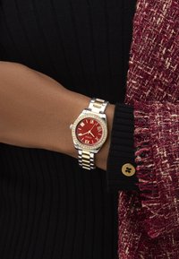 Montre en métal argenté et doré avec un cadran rouge, des chiffres romains et des accents en cristal. Portée sur un poignet avec un haut noir à côtes.