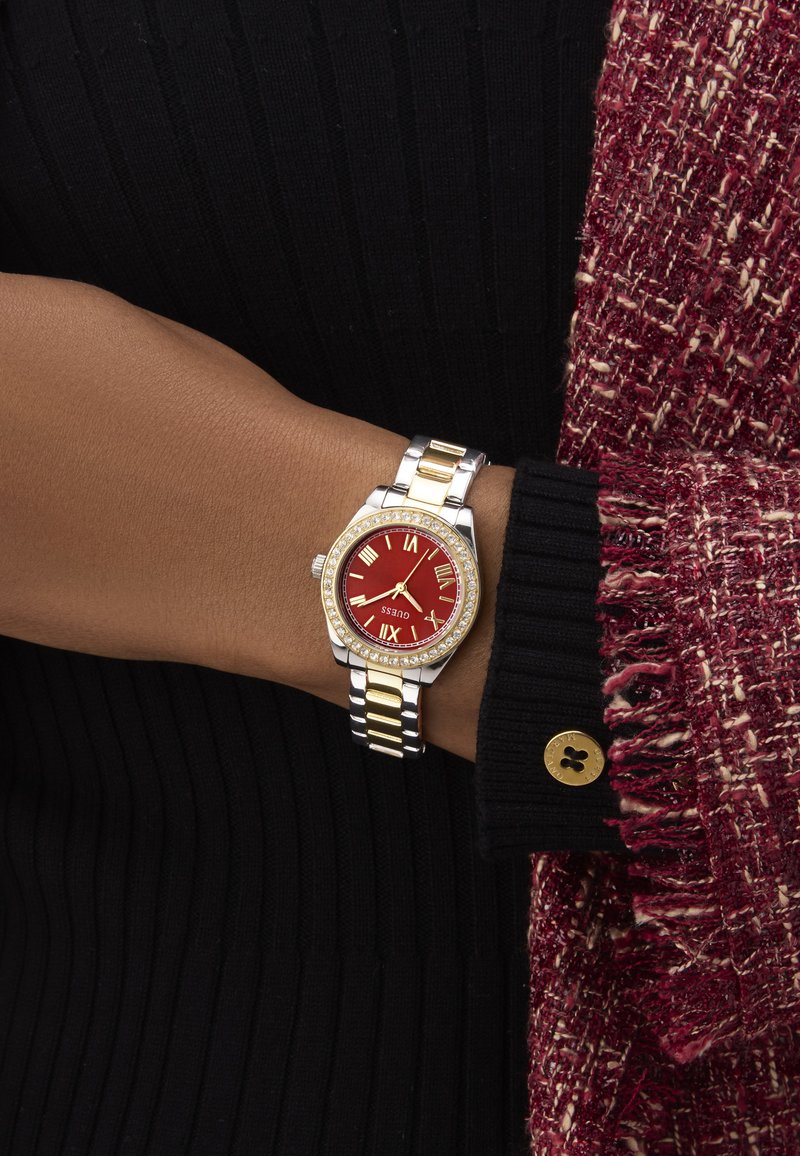 Montre en métal argenté et doré avec un cadran rouge, des chiffres romains et des accents en cristal. Portée sur un poignet avec un haut noir à côtes.