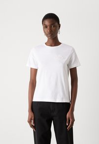 Femme portant un T-shirt blanc à manches courtes avec un logo discret sur la poitrine et un pantalon noir, debout devant un fond clair et uni.