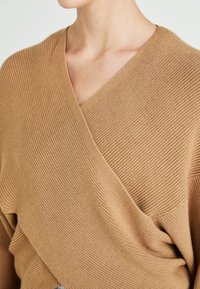 Gros plan sur une personne portant un pull cache-cœur côtelé à col en V de couleur beige avec des panneaux frontaux qui se chevauchent.