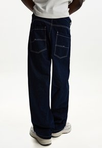 Tamno plave traperice s opuštenim krojem, s dva stražnja džepa i bijelim šavovima. Uparene s svijetlim tenisicama.