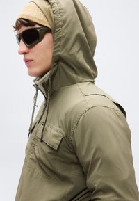 Χακί μπουφάν με κουκούλα από ελαφρύ υλικό· διαθέτει κουκούλα με κορδόνι, μπροστινή τσέπη στο στήθος και φερμουάρ. Σκούρα γυαλιά ηλίου συμπληρώνουν το ντύσιμο.