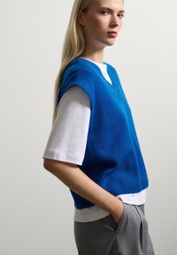 Blauwe gebreide gilet met een v-hals over een wit T-shirt, gecombineerd met grijze op maat gemaakte broek. Het gilet heeft een geribbelde zoom en brede armsgaten.
