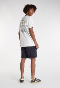 Witte katoenen t-shirt met een grafische print op de achterkant, gecombineerd met marineblauwe cargoshorts en zwart/wit gestreepte sneakers.