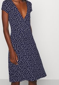 Kvinna klädd i en marinblå klänning med vita prickar, korta ärmar och djup V-ringning, som står mot en ljus och enfärgad bakgrund.