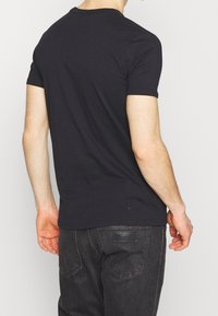 Černé bavlněné tričko s kulatým výstřihem, krátkými rukávy a hladkou strukturou, zobrazené zezadu, v kombinaci s tmavými džíny s logovanou nášivkou.