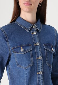 Casaco de ganga com colarinho, fecho frontal por botões e dois bolsos no peito, na cor azul média com acabamentos em metal prateado e costura amarela.