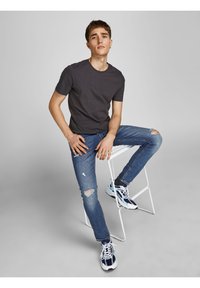 Grå bomullströja, slitna blå jeans med hål, vita och marinblå sneakers, sittande på en vit pall mot en neutral bakgrund.