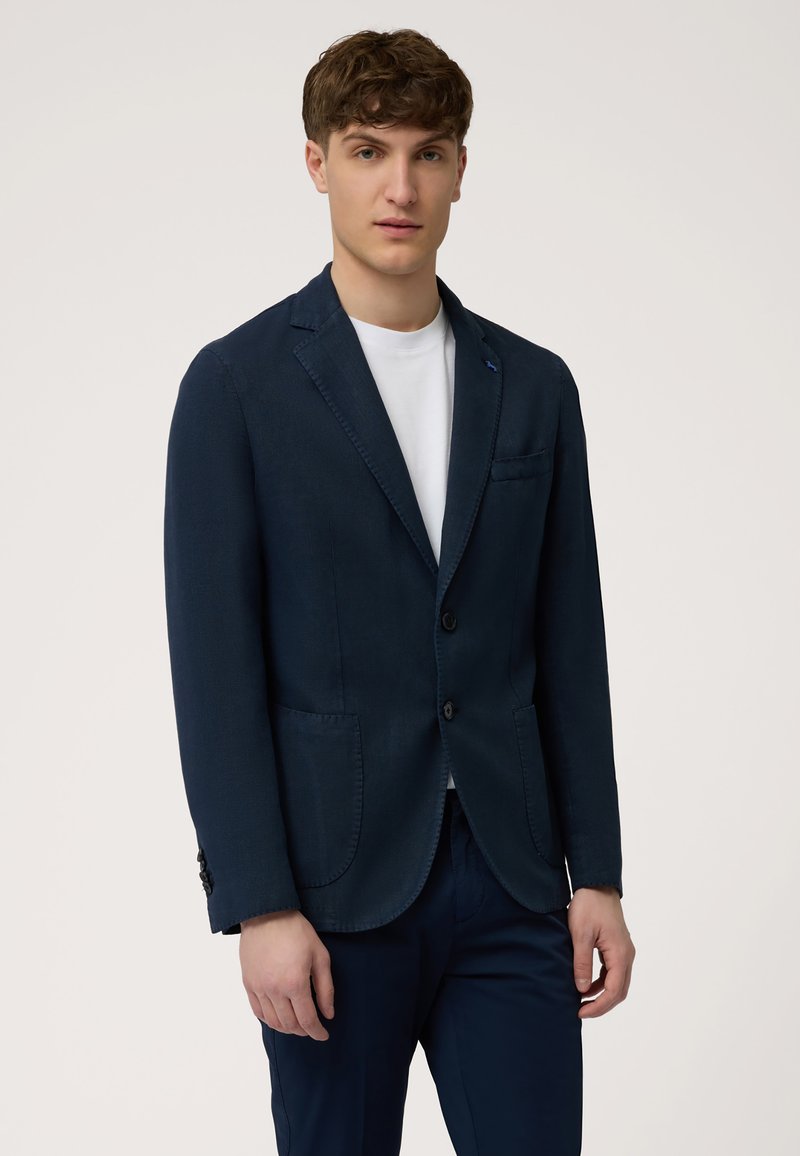 Navy blazer made of textured fabric featuring two front pockets, a single chest pocket, and black buttons. Worn over a white t-shirt.