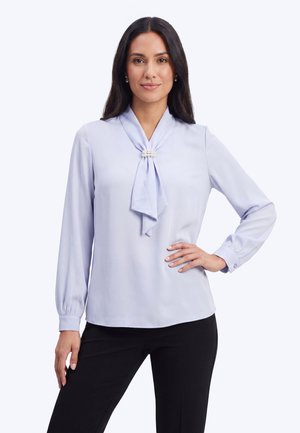 Femeie purtând o bluză albastru deschis cu mâneci lungi și o broșă cu perlă la gât și pantaloni negri, stând cu o mână pe șold.