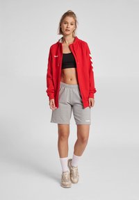 Veste zippée rouge avec des accents blancs, crop top noir et shorts gris. Détails de logo blancs ; le modèle porte des baskets beiges et des chaussettes blanches.