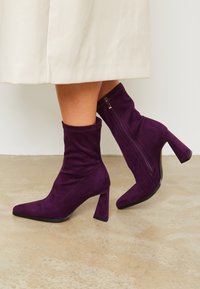 Botas de tobillo de ante púrpura con puntera afilada, diseño estilizado, tacón apilado y detalle de cremallera lateral. Suela plana con textura mínima.