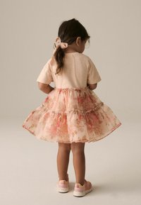 Ärmelloses, tanfarbenes Oberteil, geschichteter, pfirsichfarbener Tüllrock mit Rüschen. Das Kind trägt rosa Sneakers mit gesprenkelten Details und eine Schleife im Haar.