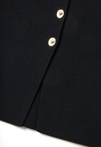 Schwarze Strickjacke mit strukturiertem Material, goldenen Knöpfen und einer leichten Öffnung am Saum. Der Stoff ist weich und glatt.