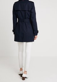 Námořnický modrý trench coat s páskem v pase, knoflíkovými akcenty, epauletami a zadním klopovým detailem, nošený přes bílé džíny a na vysokých podpatcích.