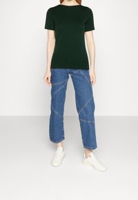 Camiseta de manga corta verde oscuro combinada con unos vaqueros cortos azules con costuras diagonales, usados con zapatillas blancas.