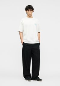 Junger Mann mit lockigem Haar, der ein weißes Kurzarm-T-Shirt mit Logo, schwarze Hosen mit weitem Bein und schwarze Turnschuhe trägt und mit den Händen in den Taschen steht.