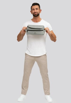 Grijze opvouwbare tas met rits, gemaakt van glad materiaal. Beschikt over een logo aan de voorkant, vastgehouden door een persoon in een wit shirt en beige broek.