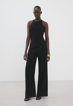 Žena u crnom širokim hlačama bez rukava s visokim ovratnikom, koja nosi zlatne narukvice i prstenje na obje ruke.