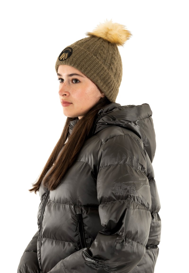 Giubbotto puffer grigio scuro con finitura lucida e cappuccio removibile, abbinato a un berretto a coste verde oliva con pompon e patch con logo.