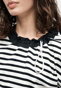 Κοντινό πλάνο ενός ατόμου που φοράει μαύρο-άσπρο ριγέ τοπ με μαύρο βολάν γύρω από το λαιμό και λευκό κορδόνι με ασημένια χάντρα.