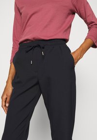 Celana hitam dengan pinggang elastis dan tali yang dapat disesuaikan, saku samping, serta tekstur yang halus, dipadukan dengan atasan maroon lengan panjang.