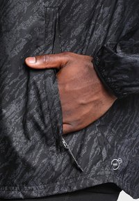 Chaqueta cortavientos negra con un patrón texturizado, bolsillos con cremallera y puños elásticos. Logo destacado en el dobladillo.