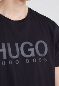 Černé bavlněné tričko s velkým šedým nápisem "HUGO" a "HUGO BOSS" na přední straně. Kulatý výstřih, krátké rukávy, hladká textura.