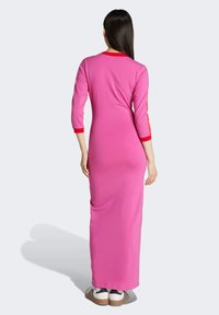 Abito lungo rosa con maniche a tre quarti e dettagli rossi sul colletto e sui polsini. Presenta una texture liscia, una silhouette aderente e tasche laterali.