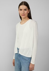 Weiße, leichte Strickjacke mit offenem Vorderteil, langen Ärmeln und weicher Textur, kombiniert mit einem weißen Oberteil und blauen Jeans. Einfaches Design.