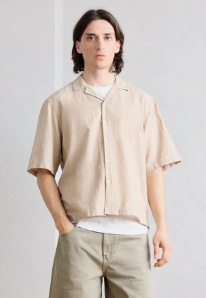 Jonge man met donker schouderlang haar, die een beige kraagloze blouse met korte mouwen draagt over een wit T-shirt en beige broek.
