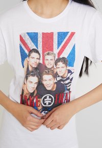 Weißes Baumwoll-T-Shirt mit einem grafischen Druck von fünf Bandmitgliedern vor einer UK-Flagge, mit dem Text "TAKE THAT" in Rot und Schwarz.