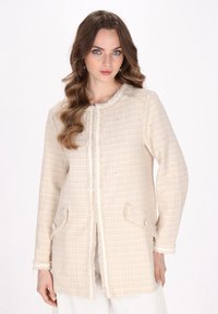 Jachetă din tweed de culoare crem, cu un model în carouri, având un tiv desfăcut, două buzunare frontale și un guler rotund.